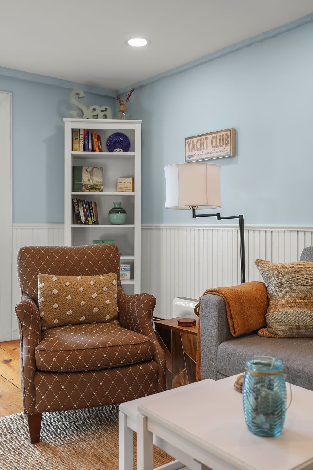 Een woonkamer met een bruine fauteuil, een witte bijzettafel, een boekenkast en een bordje met de tekst 'YACHT CLUB'.