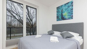 Ein Schlafzimmer mit einem großen Bett, grauer Bettwäsche, einem Pfau-Gemälde an der Wand und Blick auf Bäume durch das Fenster.