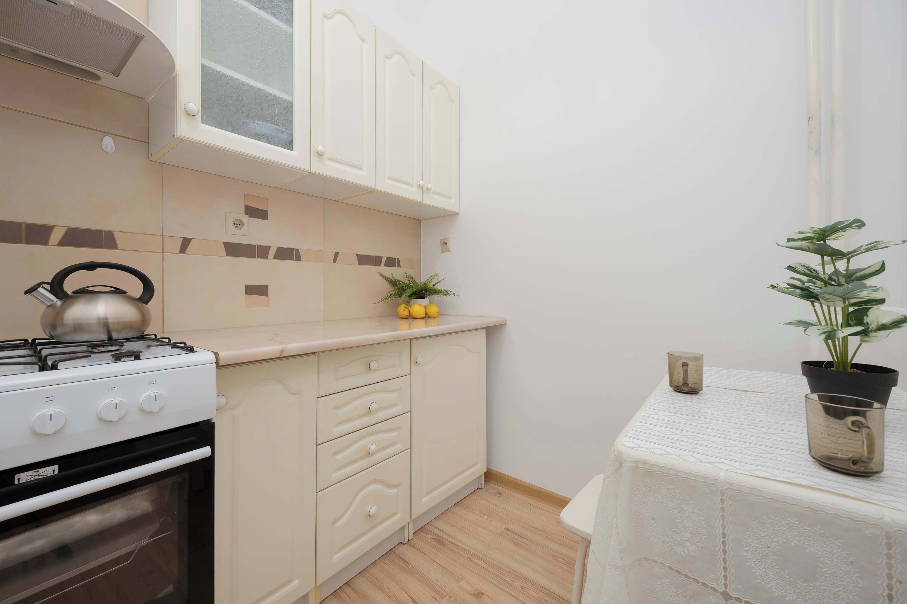 Een keuken met een fornuis, oven, kastjes, en een kleine tafel met een plant en een kop.