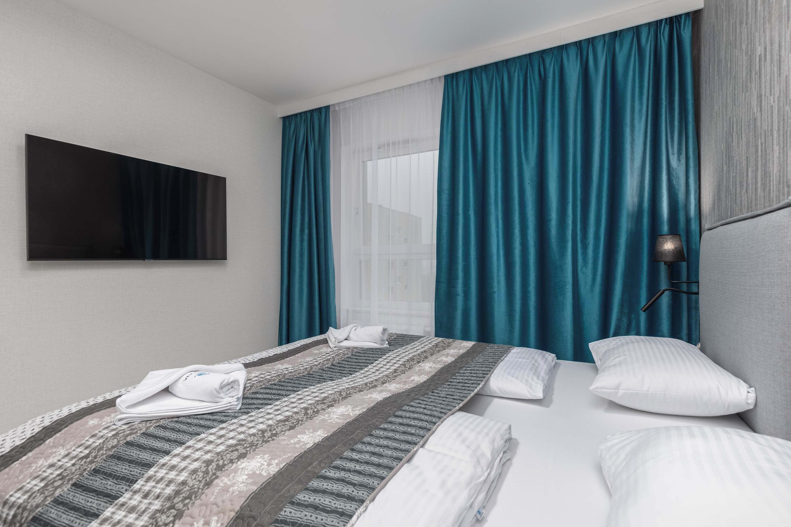 Quarto de hotel com cama, televisão fixada na parede e janela com cortinas.