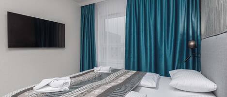 Quarto de hotel com cama, televisão fixada na parede e janela com cortinas.