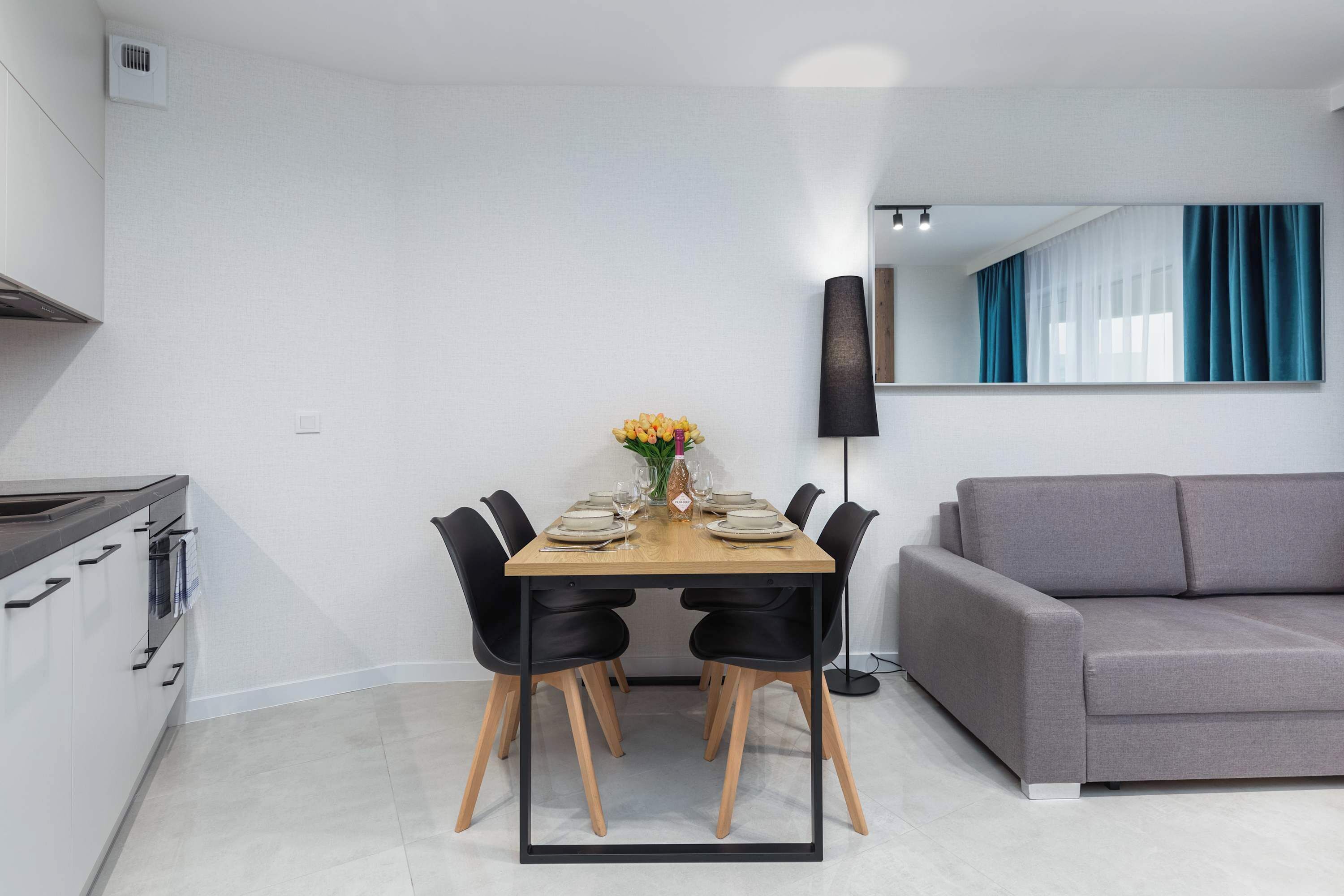 A modern dining area with a wooden table set for four, black chairs, a vase of flowers, and a grey sofa in the background.