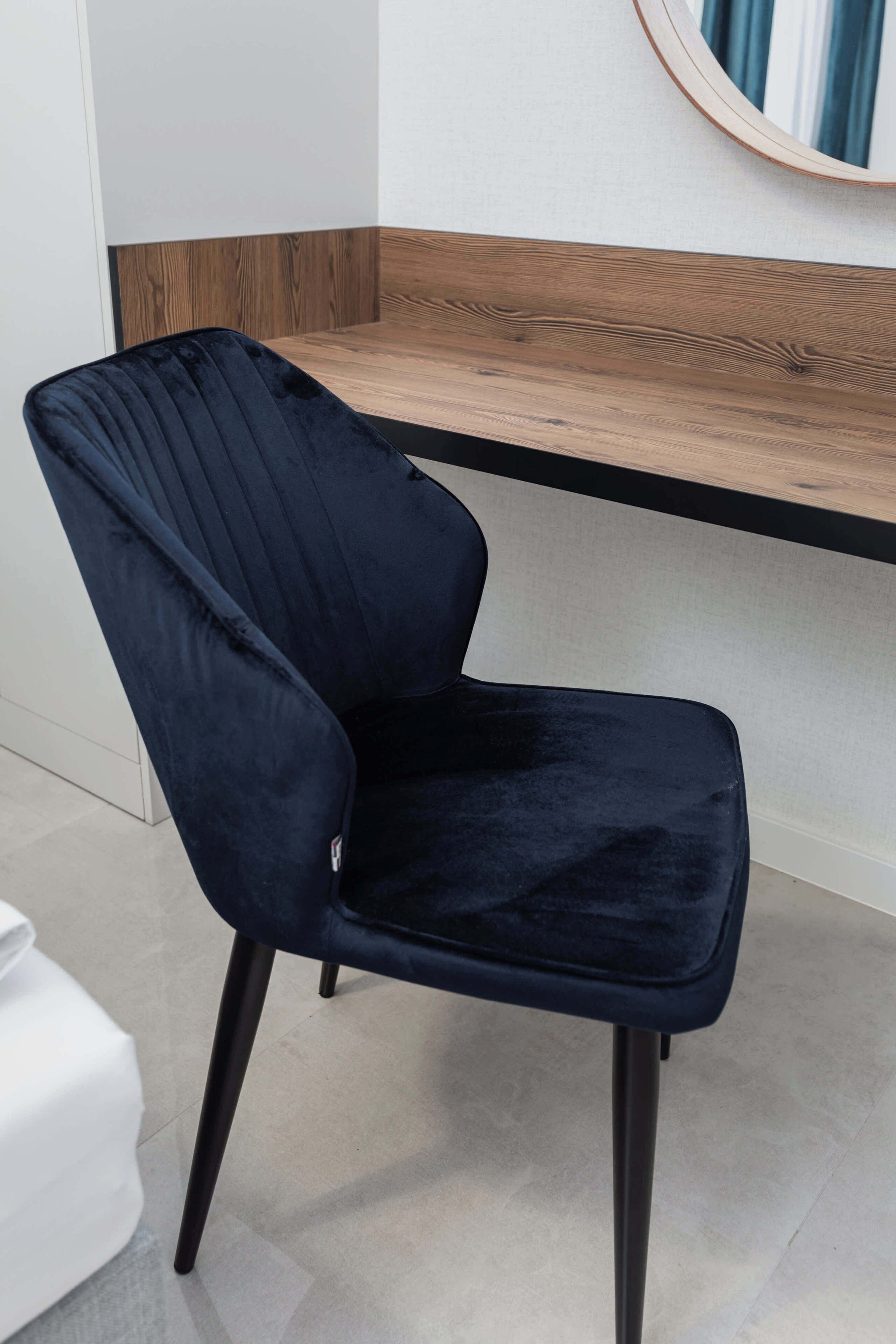 A dark blue chair with a cushioned seat and backrest, positioned in front of a wooden counter with a white wall and a round mirror above it.