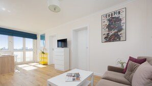 A modern living room with a sofa, a coffee table with magazines, a TV on a cabinet, and large windows with a city view.