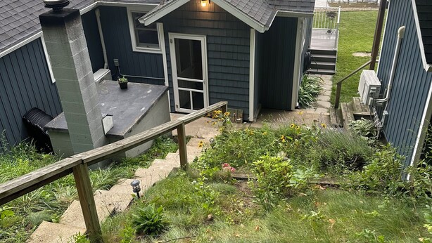 Une maison dotée d’une petite terrasse donnant sur un lac, d’une cheminée et d’un jardin agrémenté de diverses plantes.