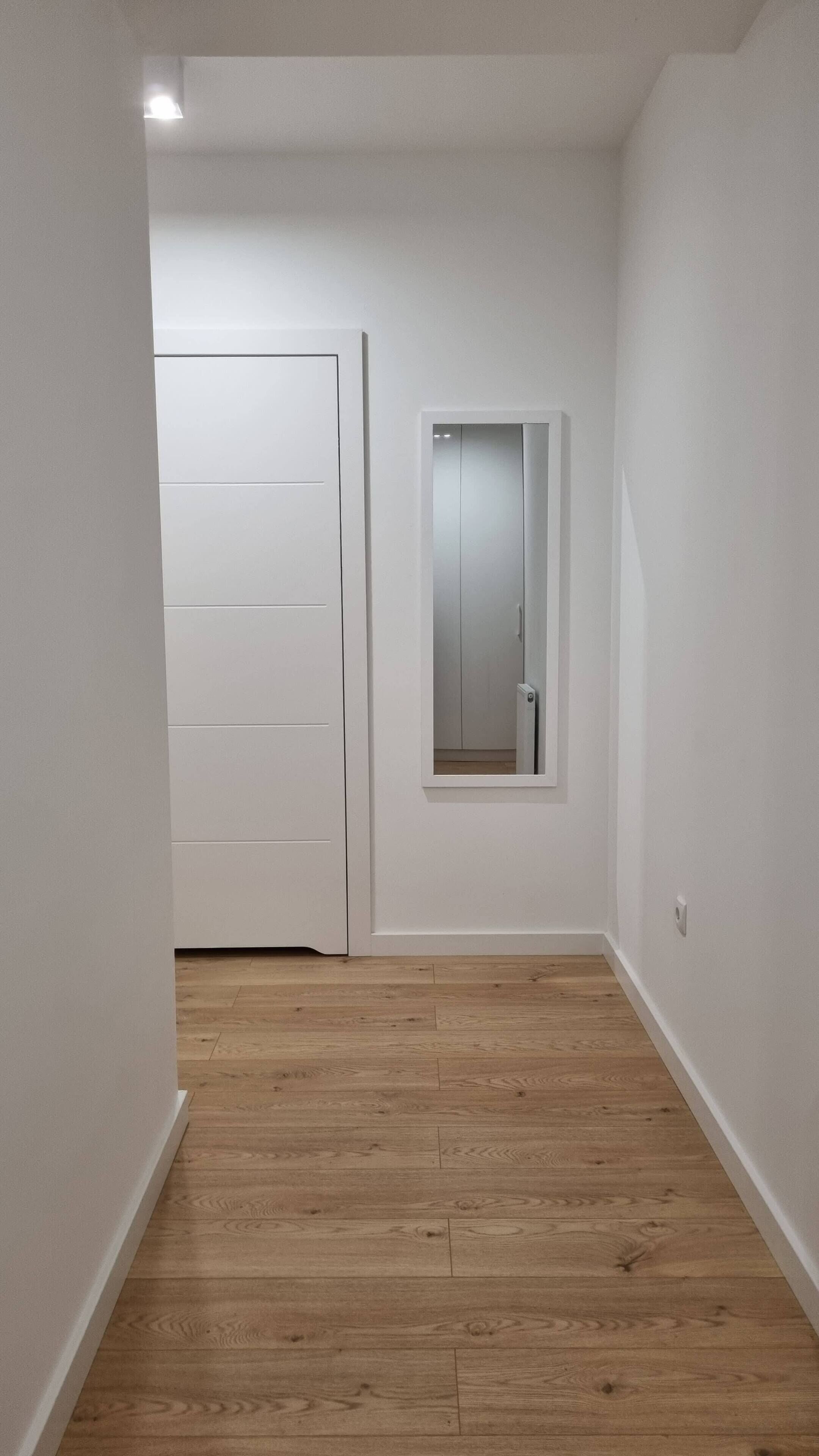 Un couloir étroit avec un plancher en bois, des murs blancs et une porte donnant sur une pièce.