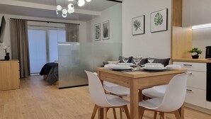 A modern dining area with a wooden table, white chairs, and a glass partition. There are framed botanical prints on the wall and a small potted plant on the counter. - Modern Studio in the Center | Balcony | Close to Market Square (Nowy Sącz)