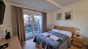 A hotel room with a bed, a nightstand, a lamp, a potted plant, and a view of the outside. - Modern Studio in the Center | Balcony | Close to Market Square (Nowy Sącz)