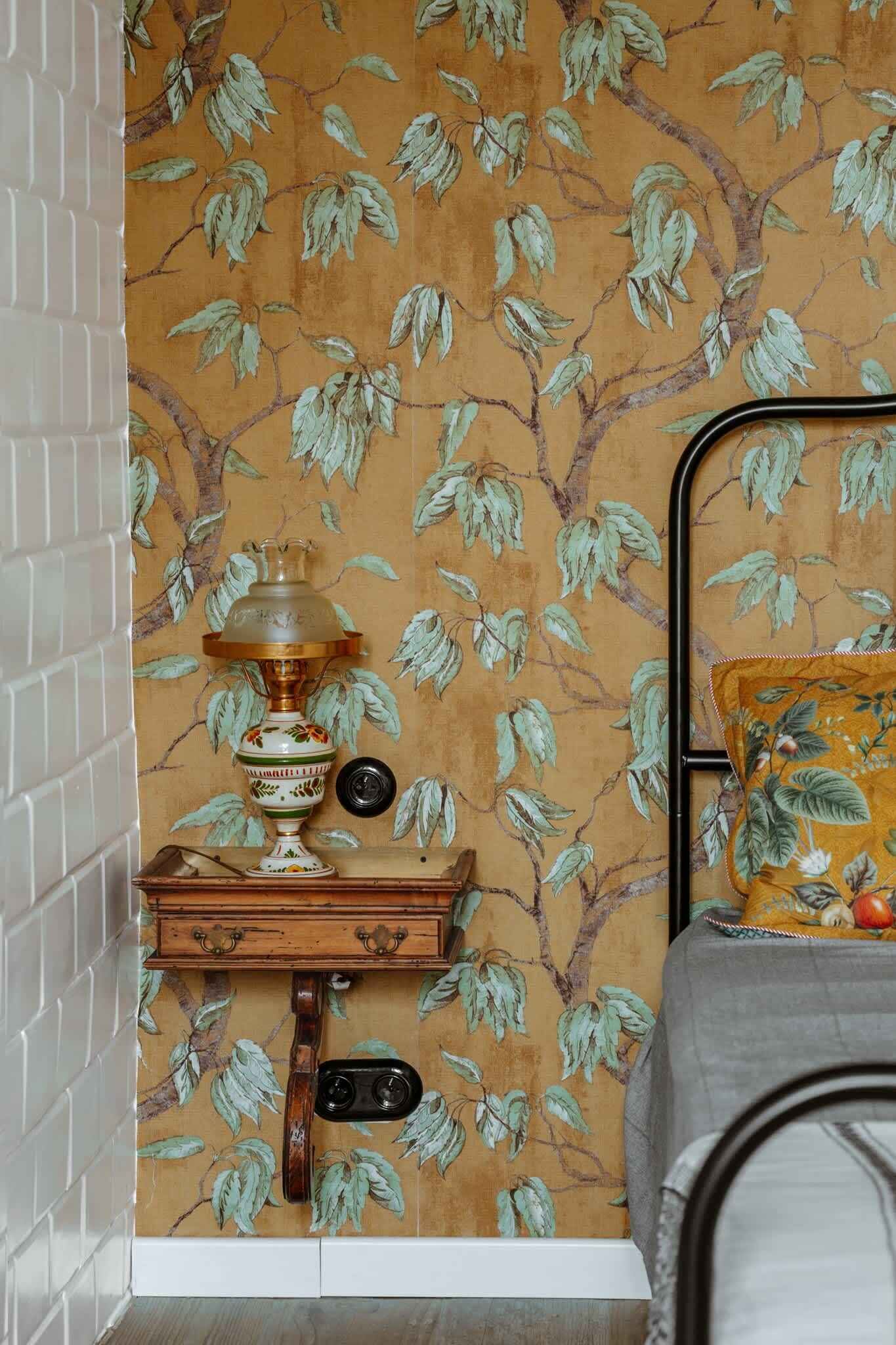 A room with a patterned wallpaper, a metal bed frame, a bedside table with a lamp, and a small wooden cabinet.