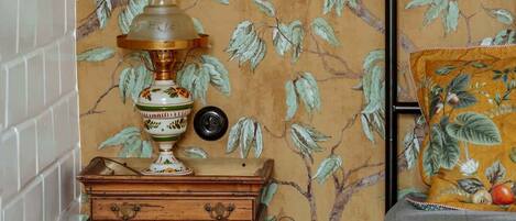 A room with a patterned wallpaper, a metal bed frame, a bedside table with a lamp, and a small wooden cabinet.