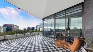 A rooftop terrace with a checkered floor, a seating area with a chair and a table, and a glass wall offering a city view.