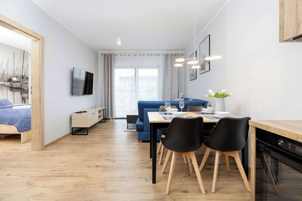 A modern living room with a sofa, dining table, and chairs. There is a TV mounted on the wall and a window with curtains.