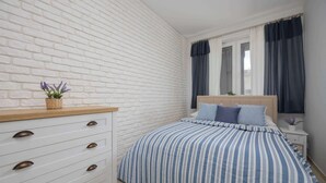 A bedroom with a white brick wall, a bed with striped bedding, a wooden dresser with drawers, and a window with curtains.