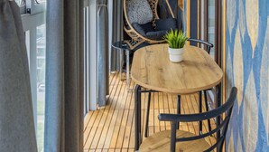 A balcony with a wooden floor, a round wooden table, two chairs, a hanging chair with cushions, and a potted plant.