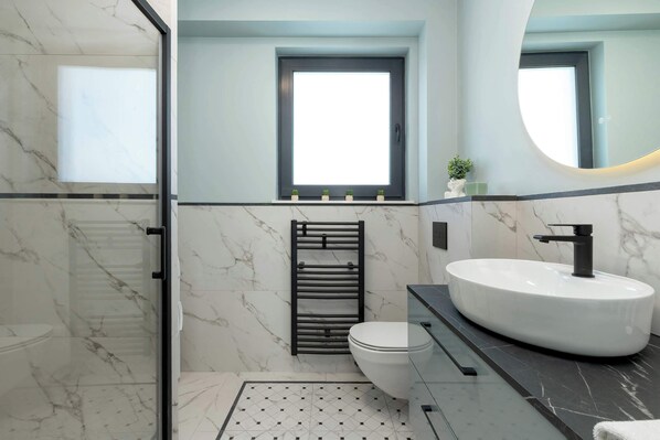 Un baño moderno con un lavamanos ovalado grande, un inodoro, un box de ducha de vidrio y una pequeña ventana sobre el lavamanos.