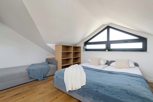 Un dormitorio moderno con techo inclinado, una cama grande con colcha azul y blanca, un sofá gris y una estantería de madera.