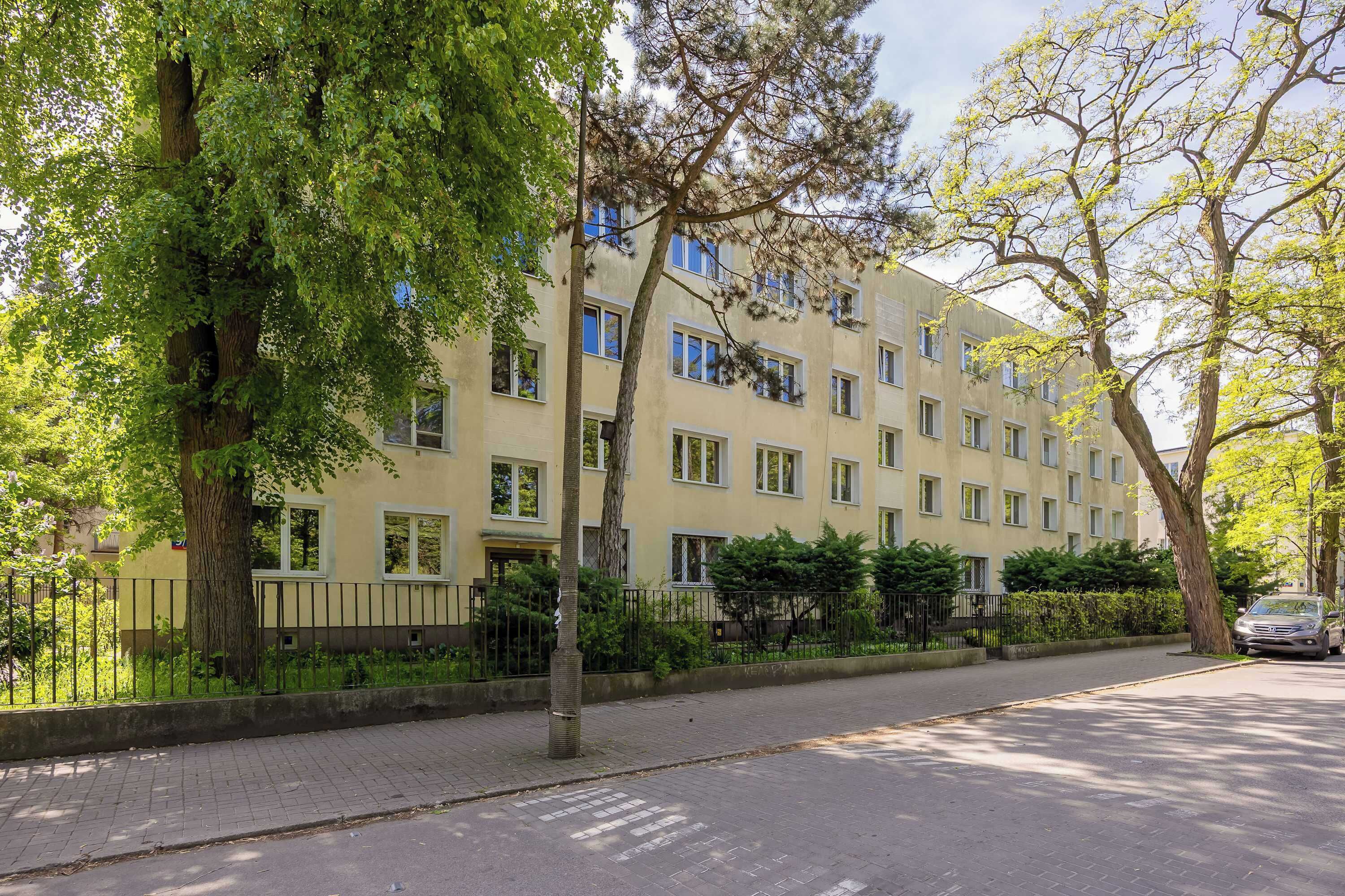 A multi-story residential building with a beige facade, multiple windows, and a paved sidewalk.