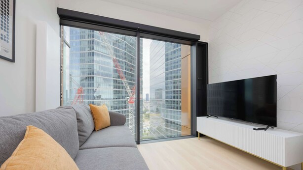 A modern living room with a grey sofa, a flat-screen TV, and a large window offering a view of a cityscape.