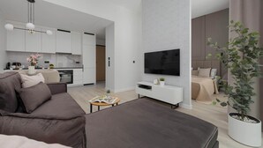 A modern living room with a sofa, a coffee table, and a TV. There is a kitchen in the background with cabinets and a dining area.