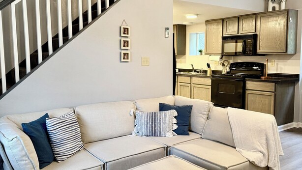 A modern living room with a sectional sofa, a small ottoman, a staircase, and a kitchen area with wooden cabinets and appliances.