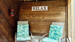 A wooden cabin interior with a 'RELAX' sign, two wicker chairs with tropical leaf cushions, a small table, and a ceiling fan.