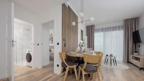 A modern living room with a dining area, wooden flooring, a flat-screen TV, and a large window with curtains.
