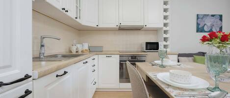 A modern kitchen with white cabinets, a stainless steel sink, and a microwave. There is a dining table set with plates, a vase with flowers, and a glass.