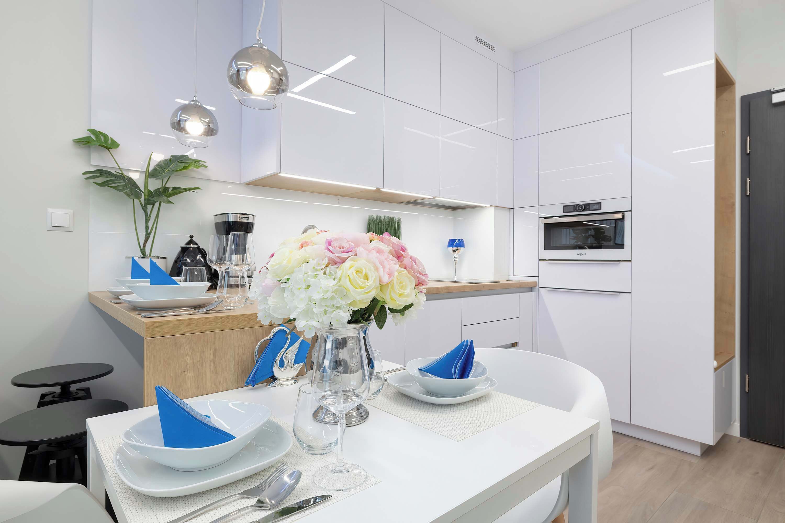A modern kitchen with white cabinets, a built-in oven, and a dining area set with white plates, blue napkins, and a vase of flowers.