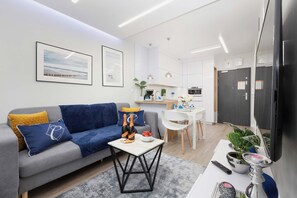 A modern living room with a grey sofa, blue and yellow cushions, a small table with a bottle and flowers, and a kitchen in the background.
