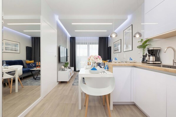 A modern kitchen with white cabinetry, a dining area with a white table and chairs, and a living room with a sofa and a TV.