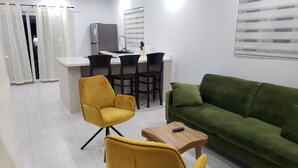 Een knus woongedeelte met een kitchenette, een groene bank en twee gele stoelen.