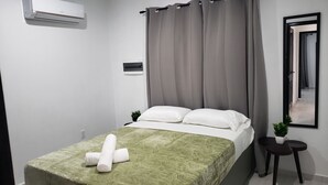 Een keurig opgemaakt bed met een groene sprei en witte kussens, een klein nachtkastje met een plantje, en een raam met grijze gordijnen.