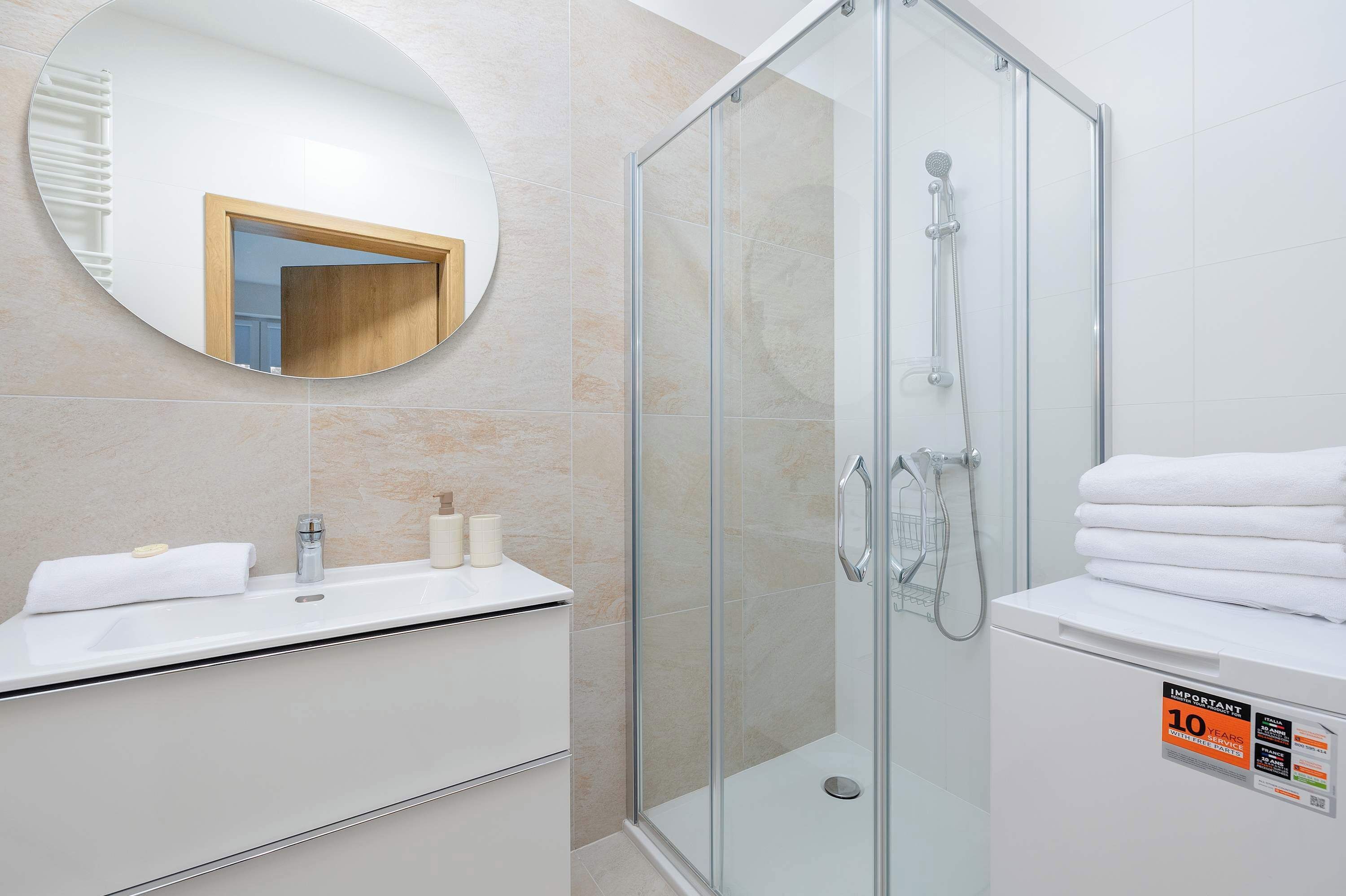 A modern bathroom with a glass shower enclosure, a round mirror, a white sink, and a toilet with a towel stack.