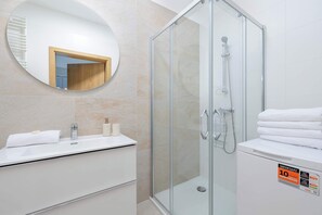 A modern bathroom with a glass shower enclosure, a round mirror, a white sink, and a toilet with a towel stack.
