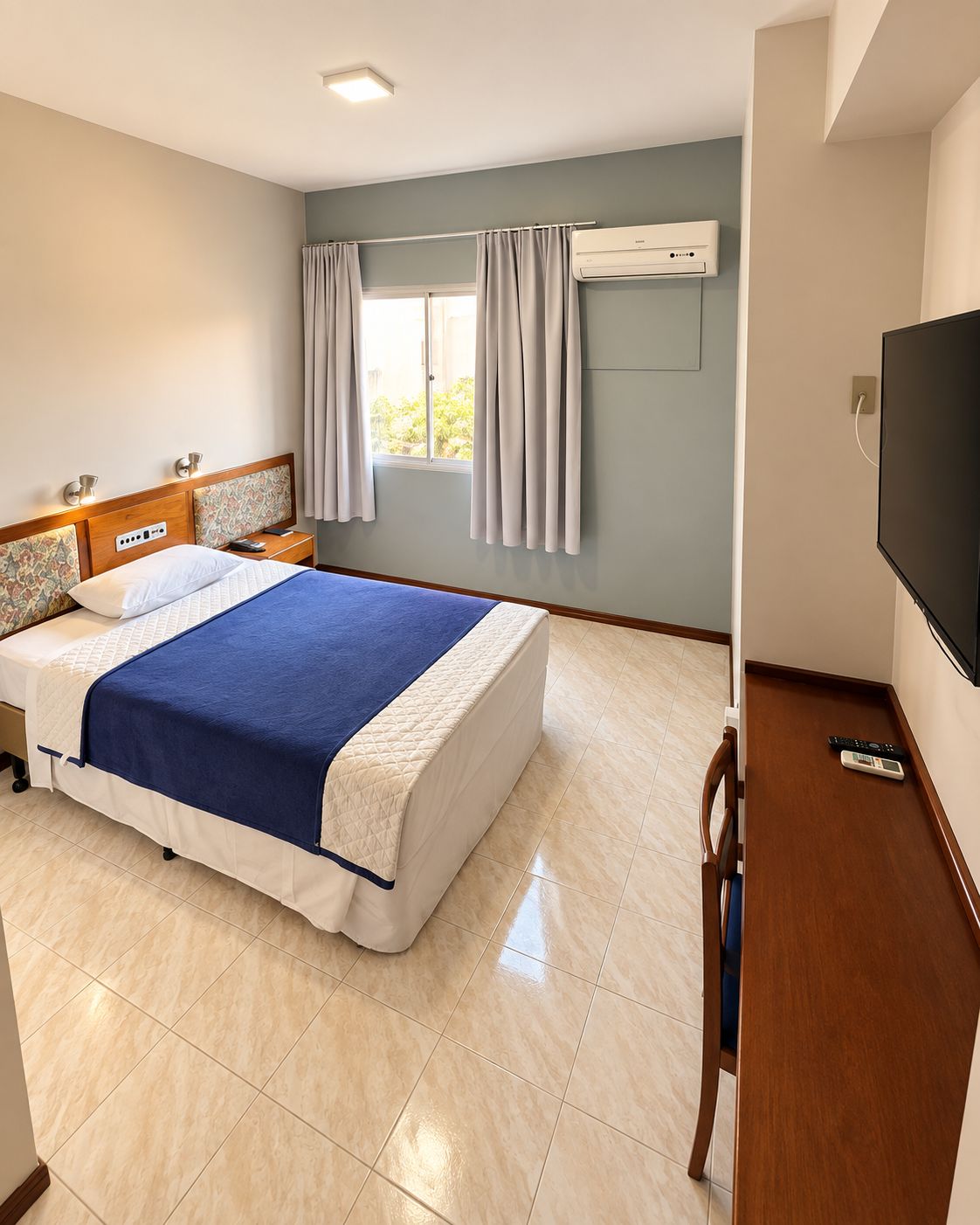 A hotel room with a bed, bedside tables, a TV, and an air conditioning unit.
