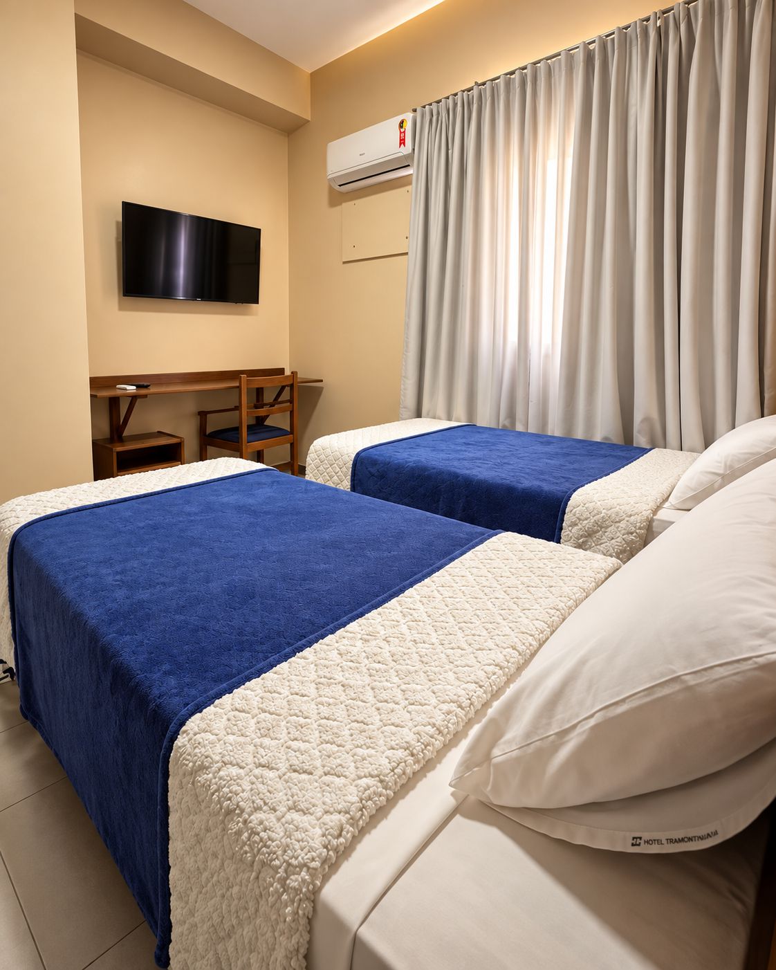 A hotel room with two beds, a desk, a chair, a television, and an air conditioning unit.