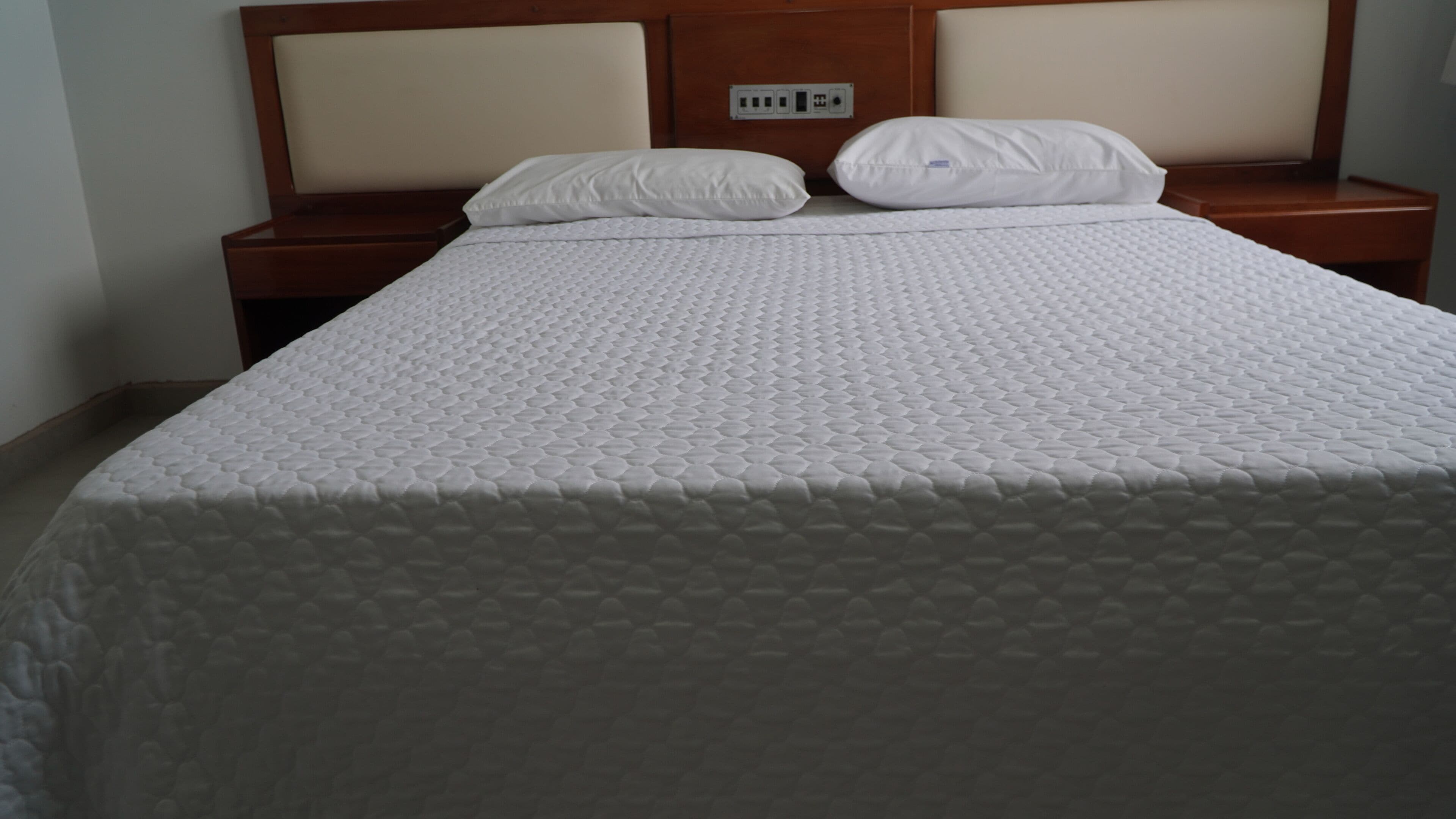 A neatly made bed with a quilted mattress and two pillows, a wooden headboard, and a bedside table with a power outlet.