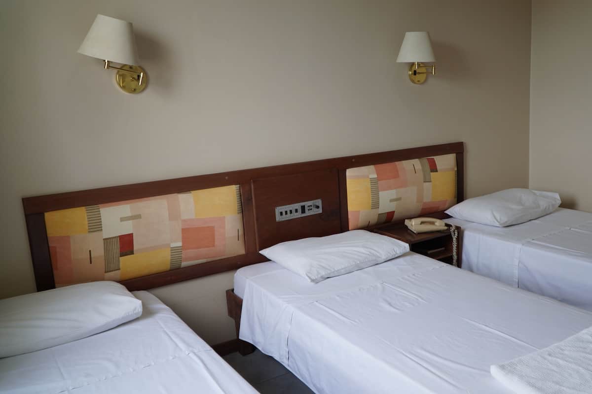 A hotel room with three single beds, a wooden headboard with a geometric pattern, a bedside table with a telephone, and two wall-mounted lamps.