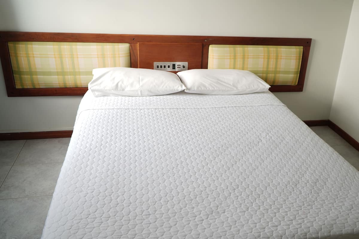 A neatly made bed with a quilted mattress, two pillows, and a wooden headboard with a built-in light switch.