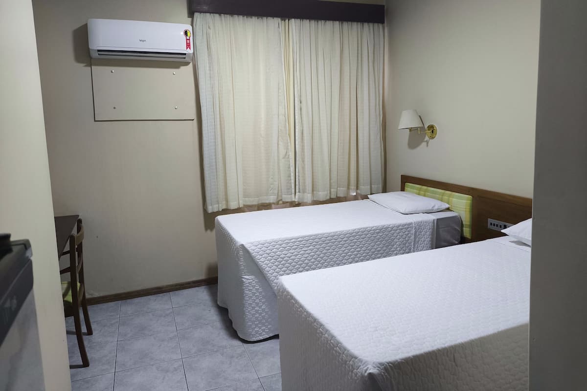 A hotel room with two beds, a window with curtains, an air conditioning unit, and a wall-mounted lamp.
