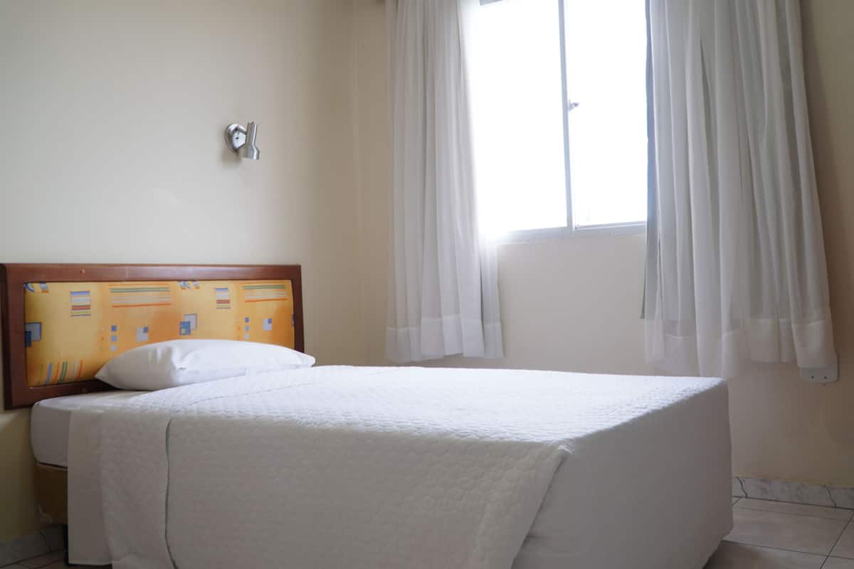 A hotel room with a bed, a window with curtains, a wooden headboard, and a wall-mounted light switch.