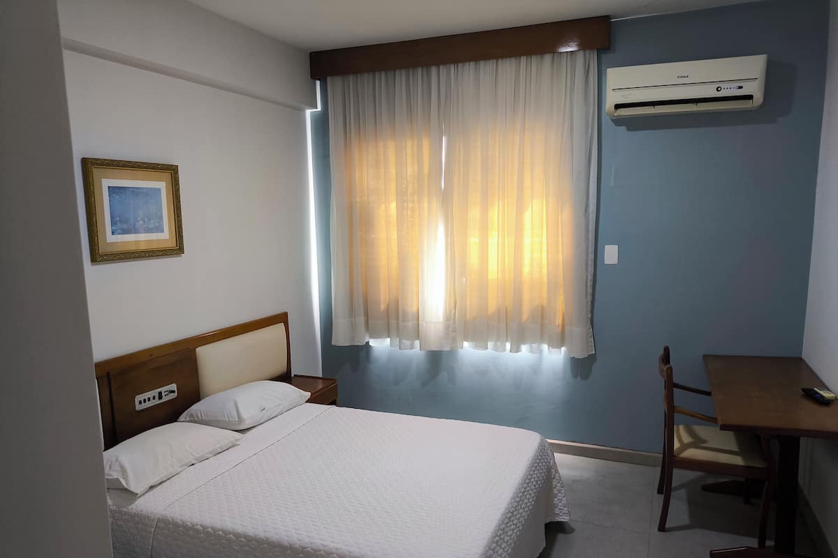 A hotel room with a bed, a desk, and an air conditioning unit.