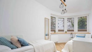 A modern bedroom with a white bed, a white sofa, a large mirror, and a view of trees through the windows.