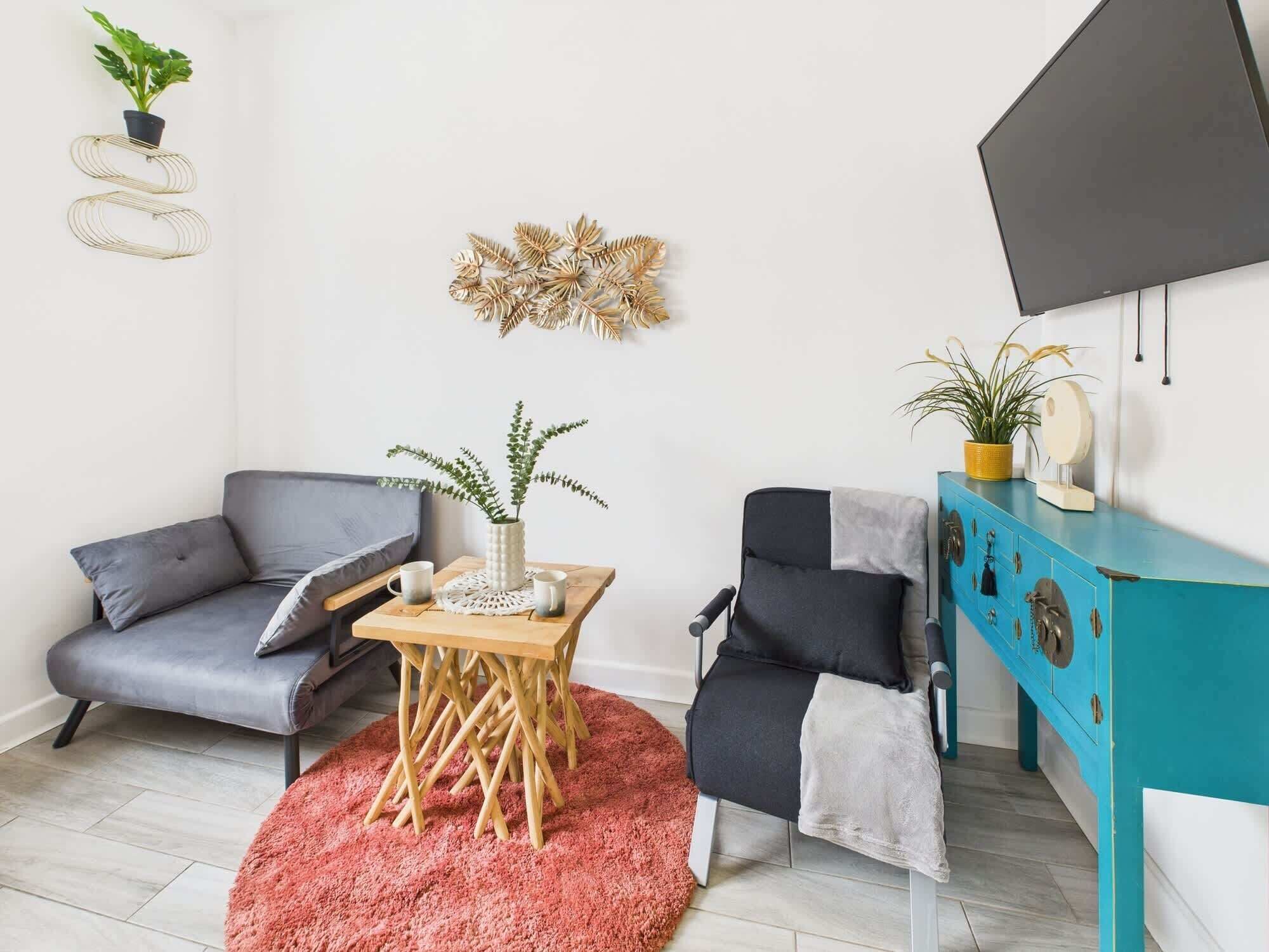 A modern living room with a grey sofa, a black and white armchair, a wooden coffee table with a vase of green plants, a pink rug, and a teal cabinet with decorative items.