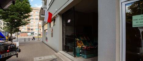 Vista de la calle con un edificio que tiene una bandera y un letrero que indica un negocio. Hay cajas afuera del edificio, y un coche estacionado con una matrícula visible.