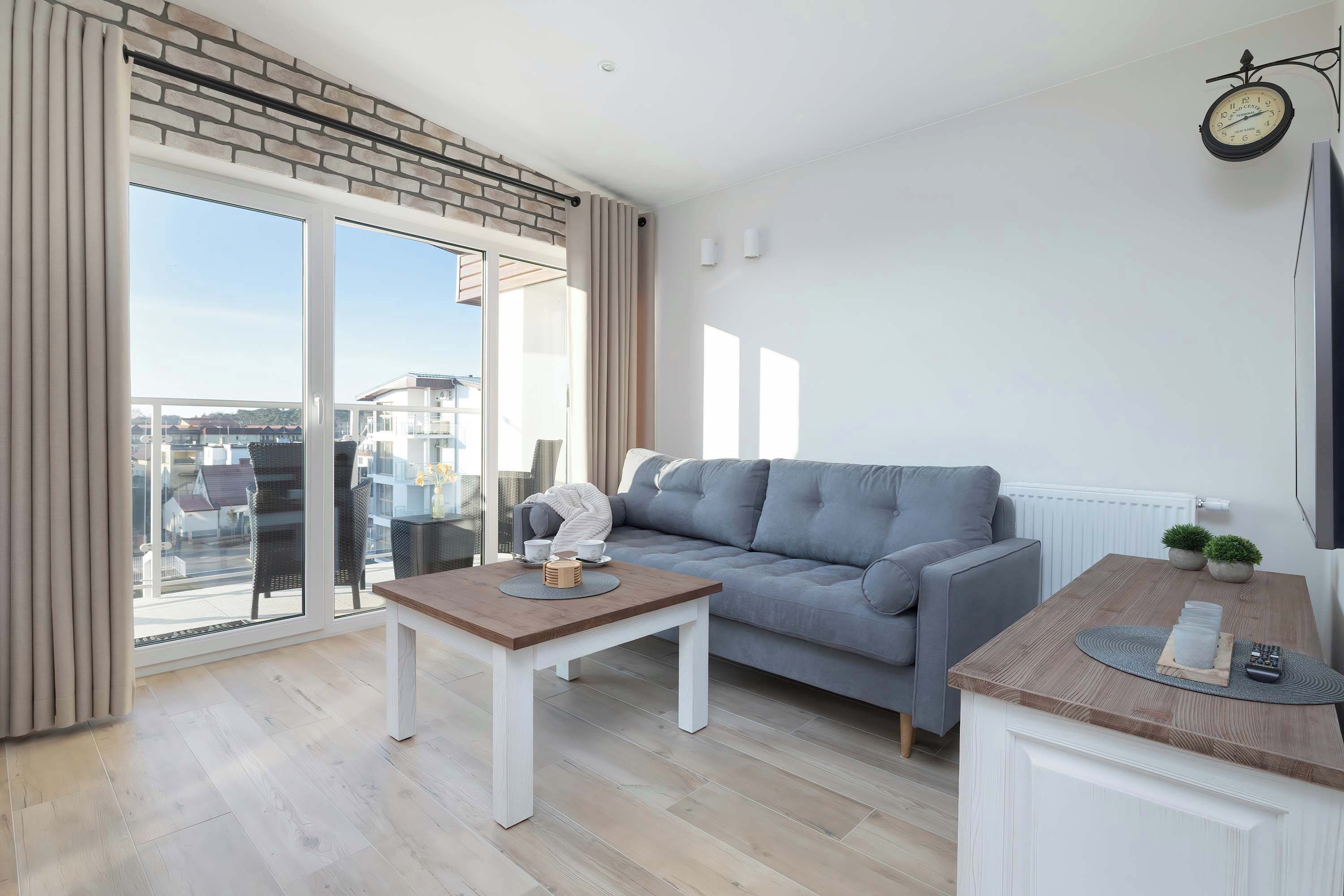 A modern living room with a grey sofa, a wooden coffee table, and a small kitchen area with a counter and a clock on the wall.