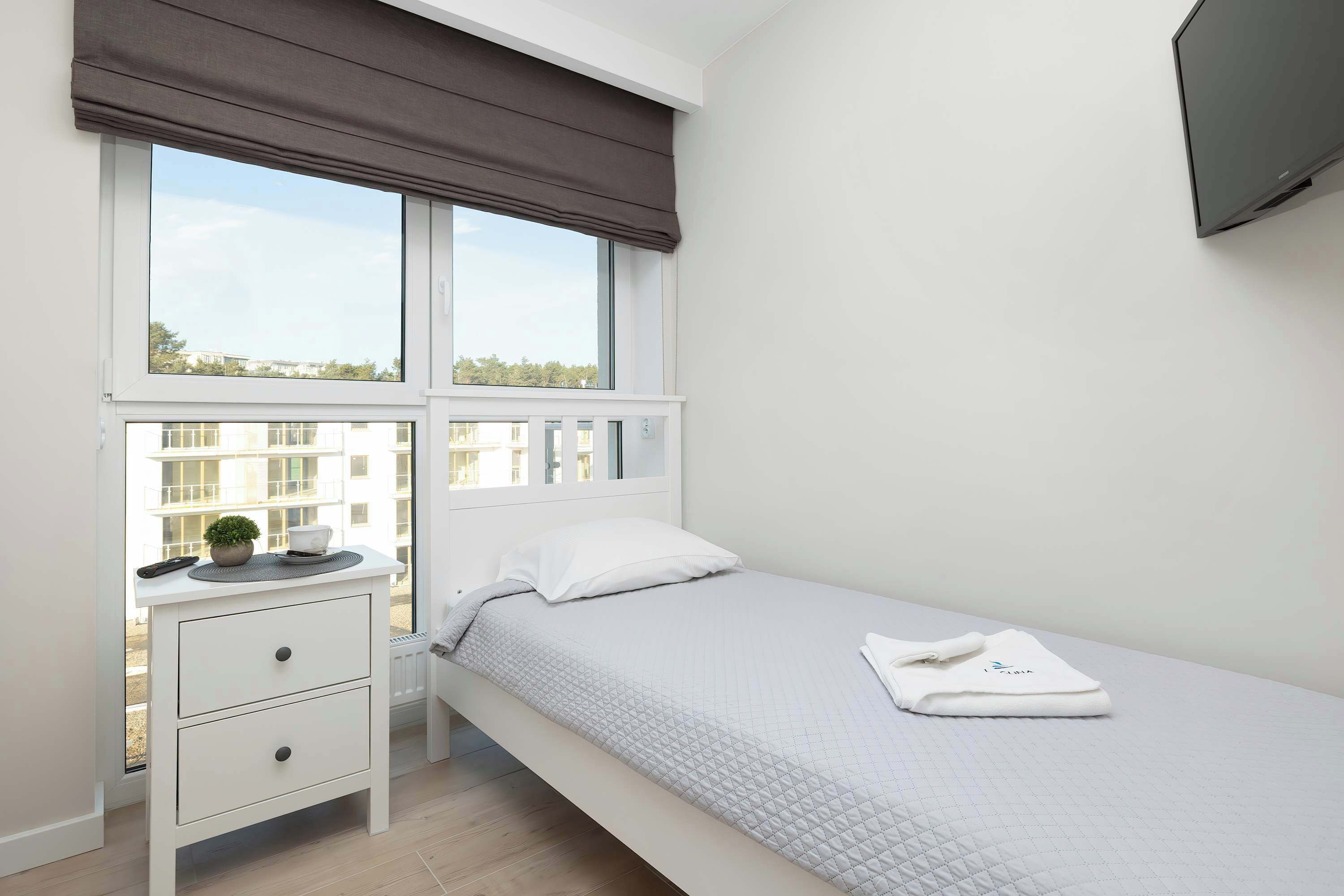 Un dormitorio con una cama, una mesita de noche, una ventana con vistas a edificios y un televisor montado en la pared.