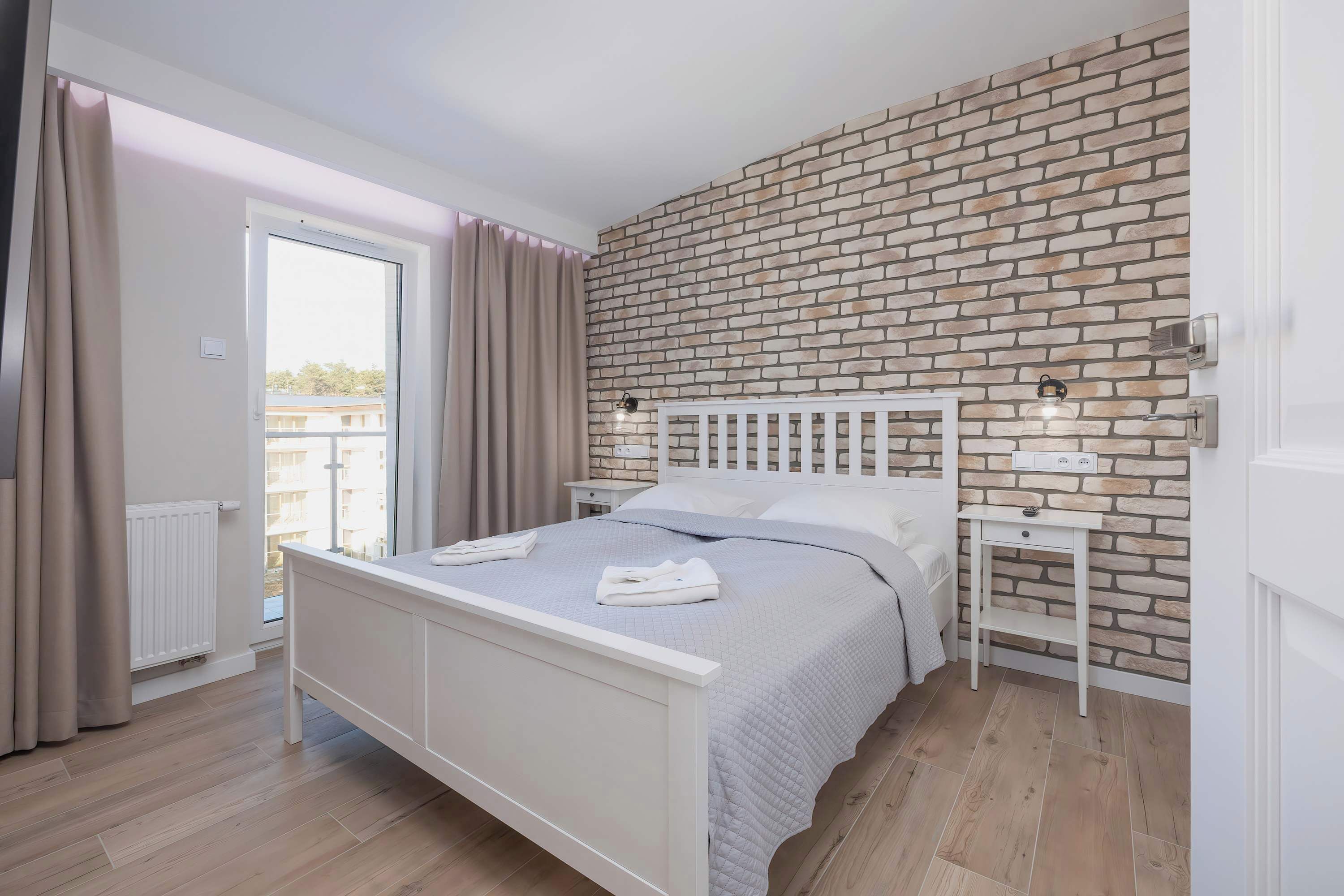 A bedroom with a white bed, a brick-patterned wall, a window with curtains, and a nightstand with a lamp.