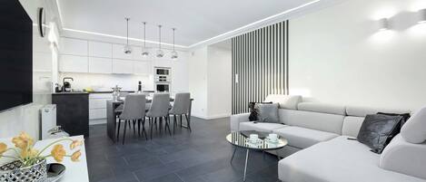 A modern living room with a white sofa, a coffee table, and a dining area with chairs and a kitchen in the background.