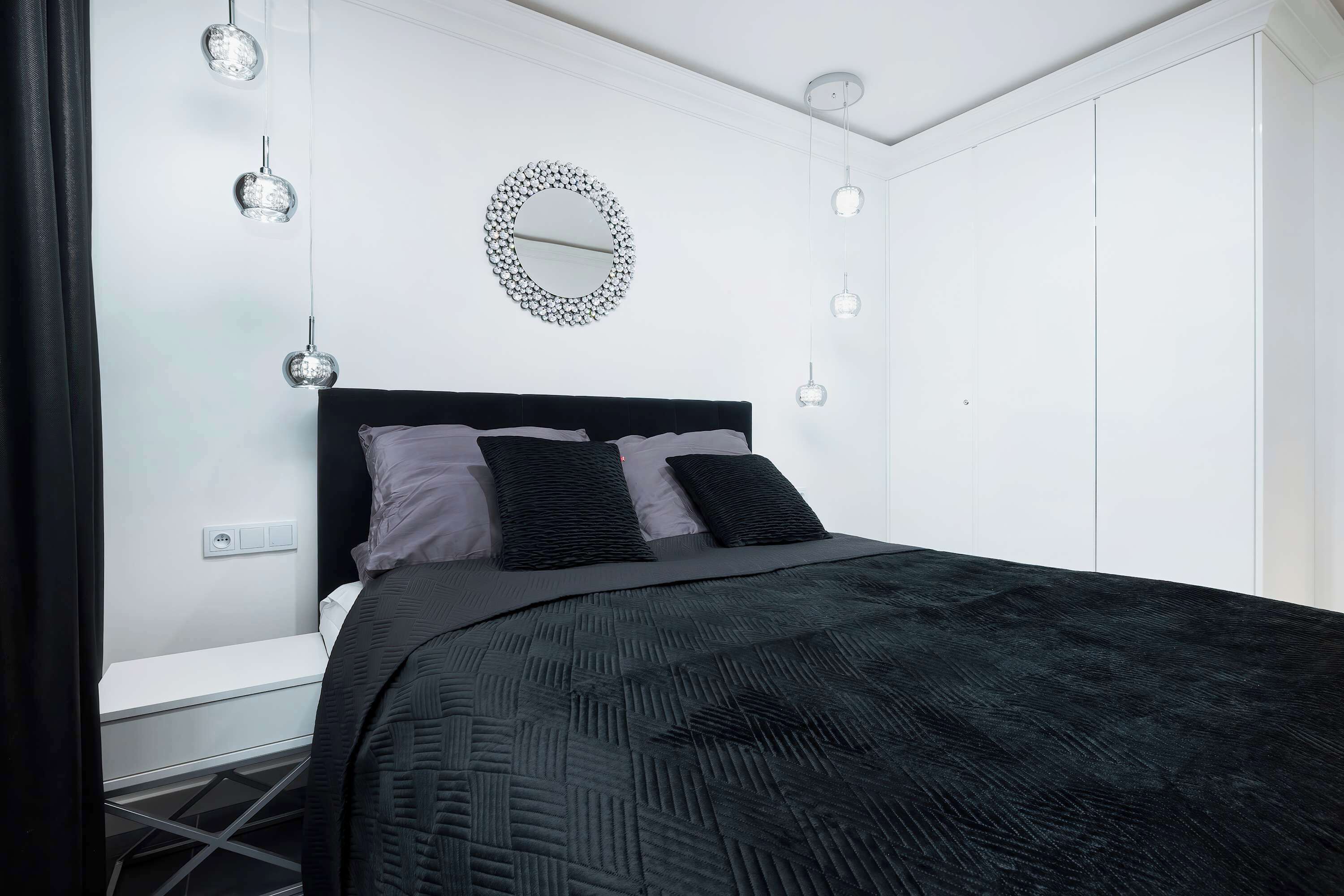 A bedroom with a black bedspread, a round mirror on the wall, and pendant lights.